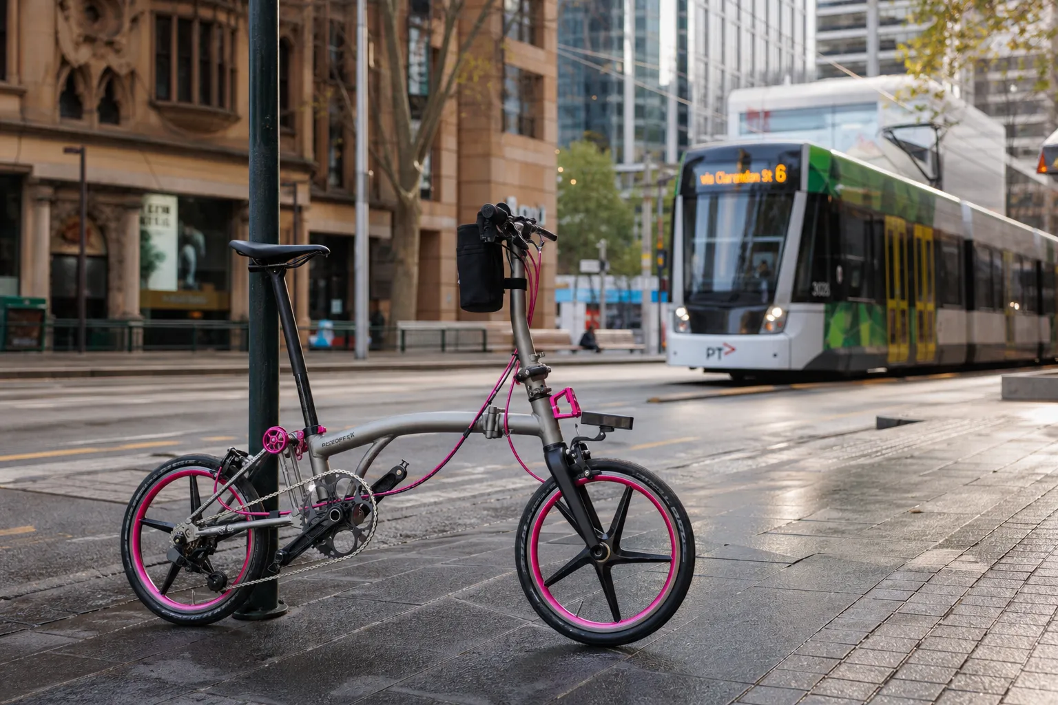 A folding bike with pink wheel accents parked on a Melbourne street, with a tram passing in the background