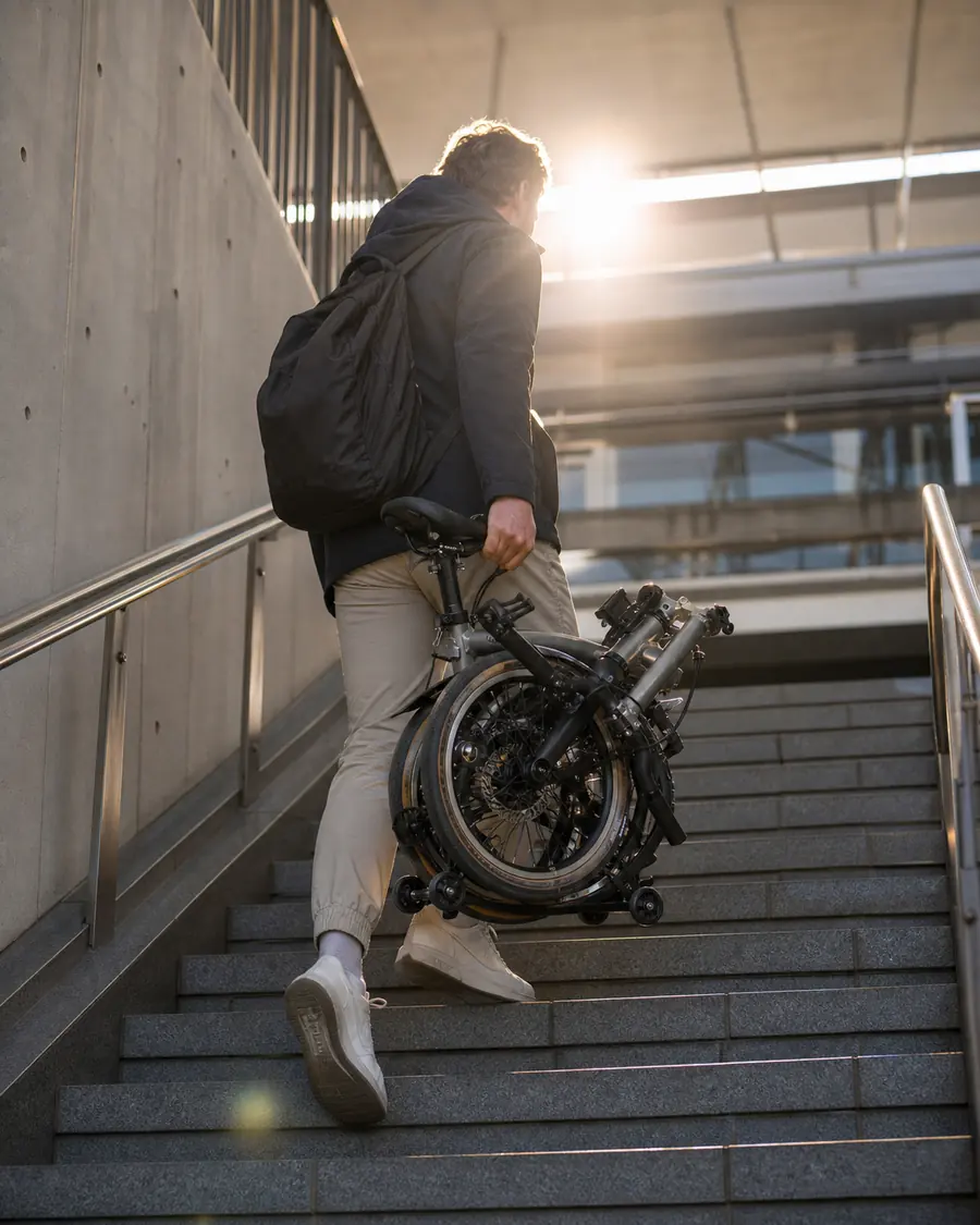 A commuter carries a folded LMile bike up a station stairwell with the morning sun behind them.